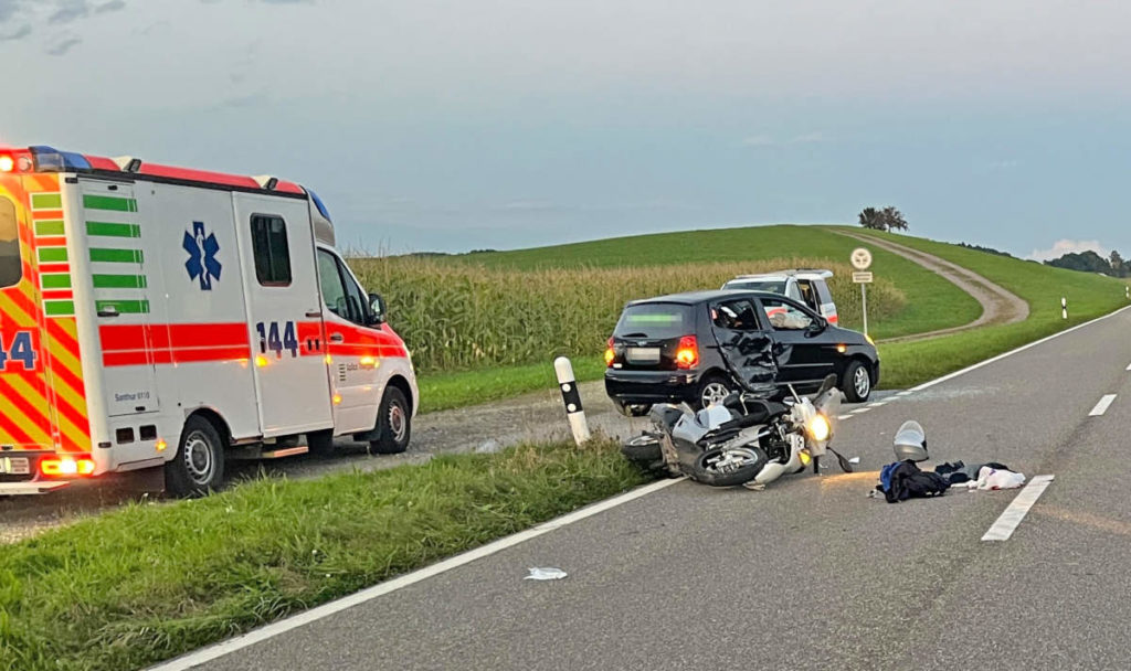 Beschädigter Roller liegt auf Strasse, am Strassenrand das an der Seite ebenfalls beschädigte Auto. Dahinter steht auf dem Grünstreifen am Maisfeld ein Rettungswagen.