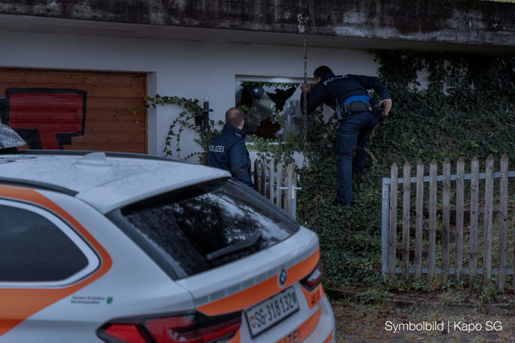 Symbolfoto: Polizeiauto vor Haus, ein Polizist steht vorm Gartenzaun, einer steht im Gebüsch vor einem Fenster und leuchtet hinein