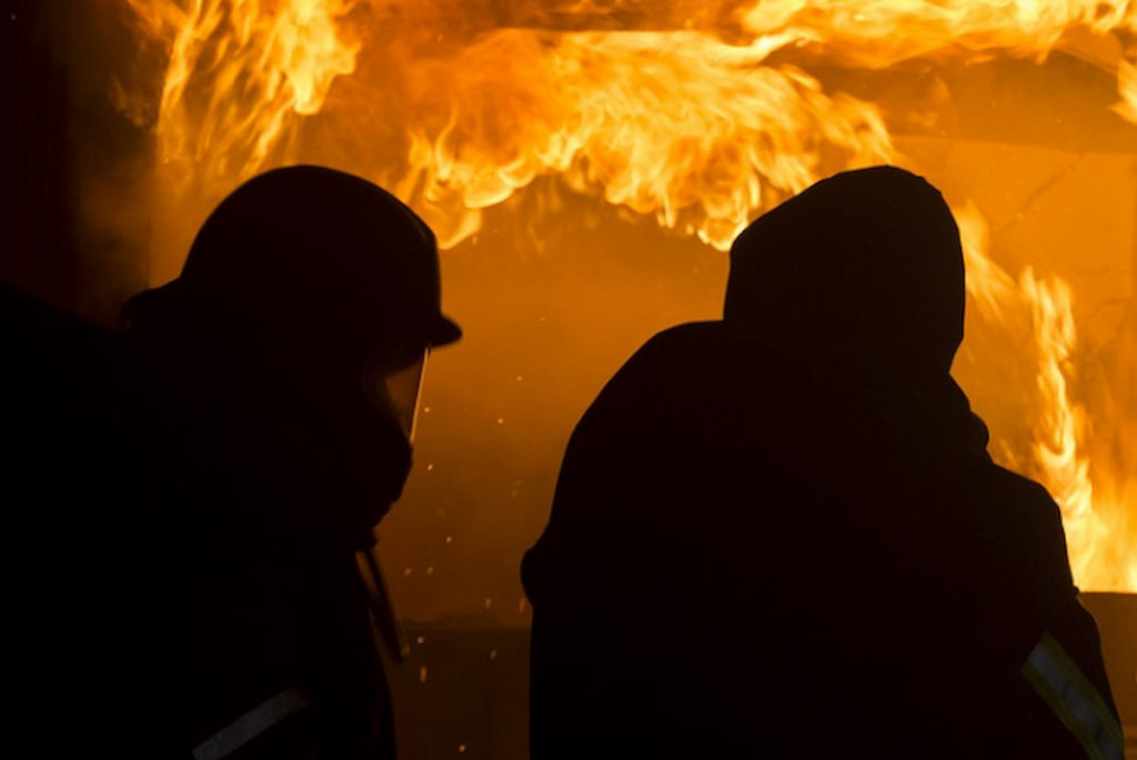 Symbolfoto: zwei als Silhouetten zu sehende Feuerwehrkräfte vor Feuerwand