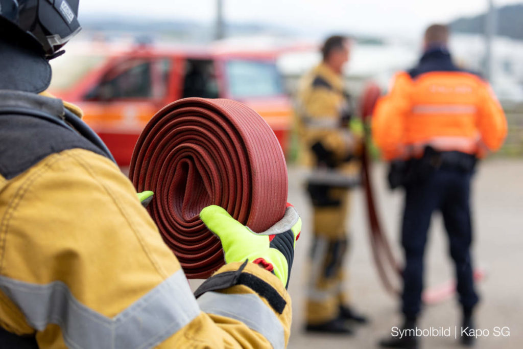 Symbolfoto: Feuerwehrkraft hält aufgerollten Schlauch in der Hand, im Hintergrund weitere Einsatzkräfte